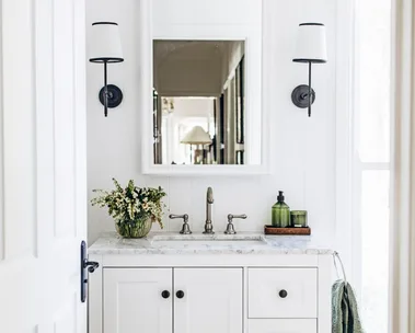 A white country bathroom with green decorative details such as rolled green towels and green hand soap. Two lamps are positioned on either side of the arched mirror, above a freestanding vanity with shaker cabinets and a marble top.
