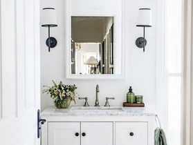 A white country bathroom with green decorative details such as rolled green towels and green hand soap. Two lamps are positioned on either side of the arched mirror, above a freestanding vanity with shaker cabinets and a marble top.
