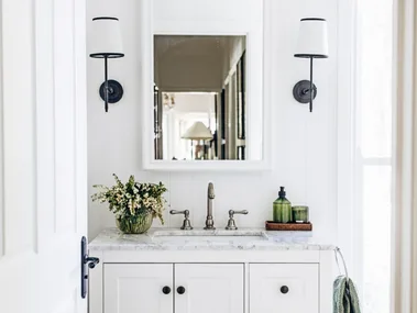 A white country bathroom with green decorative details such as rolled green towels and green hand soap. Two lamps are positioned on either side of the arched mirror, above a freestanding vanity with shaker cabinets and a marble top.