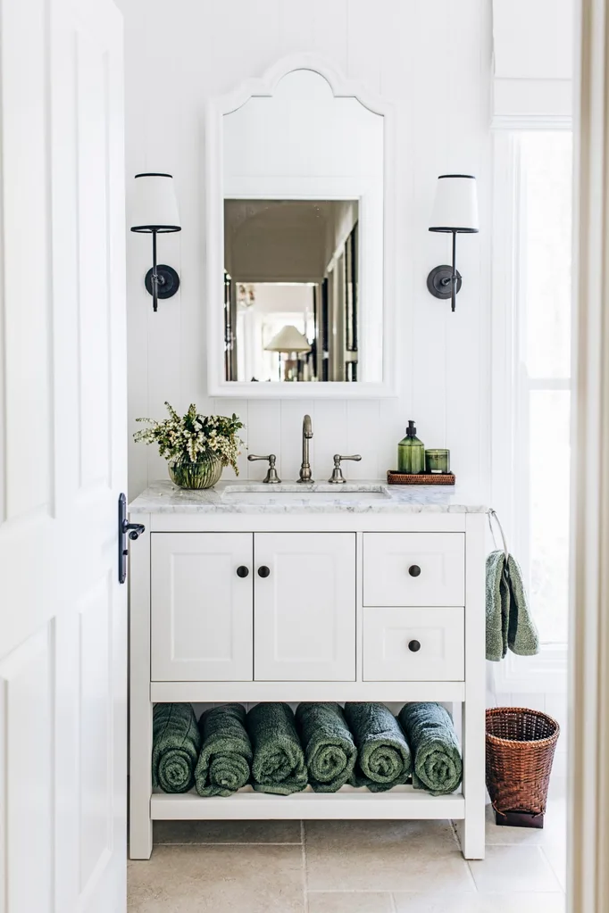 A white country bathroom with green decorative details such as rolled green towels and green hand soap. Two lamps are positioned on either side of the arched mirror, above a freestanding vanity with shaker cabinets and a marble top.