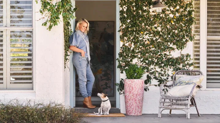 Woman in denim stands in doorway with a small dog. Vines decorate the house exterior; a wicker chair sits nearby.