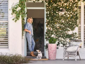 Woman in denim stands in doorway with a small dog. Vines decorate the house exterior; a wicker chair sits nearby.