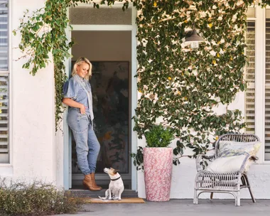 Woman in denim stands in doorway with a small dog. Vines decorate the house exterior; a wicker chair sits nearby.