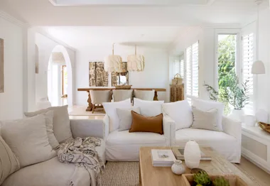 Cozy living room with neutral tones, white sofas, wooden table, and plants near large windows with shutters.