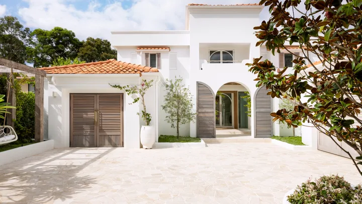Mediterranean style white house with terracotta roof, wooden shutters, and a stone driveway, surrounded by greenery.