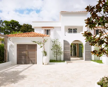 Mediterranean style white house with terracotta roof, wooden shutters, and a stone driveway, surrounded by greenery.