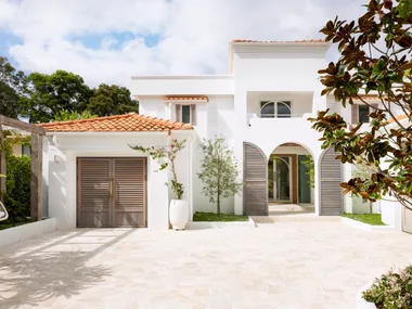 Mediterranean style white house with terracotta roof, wooden shutters, and a stone driveway, surrounded by greenery.
