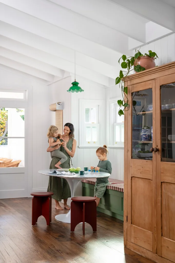 A mother with two children in a bright kitchen, with wooden floors, bench seating, and a table with green decor.