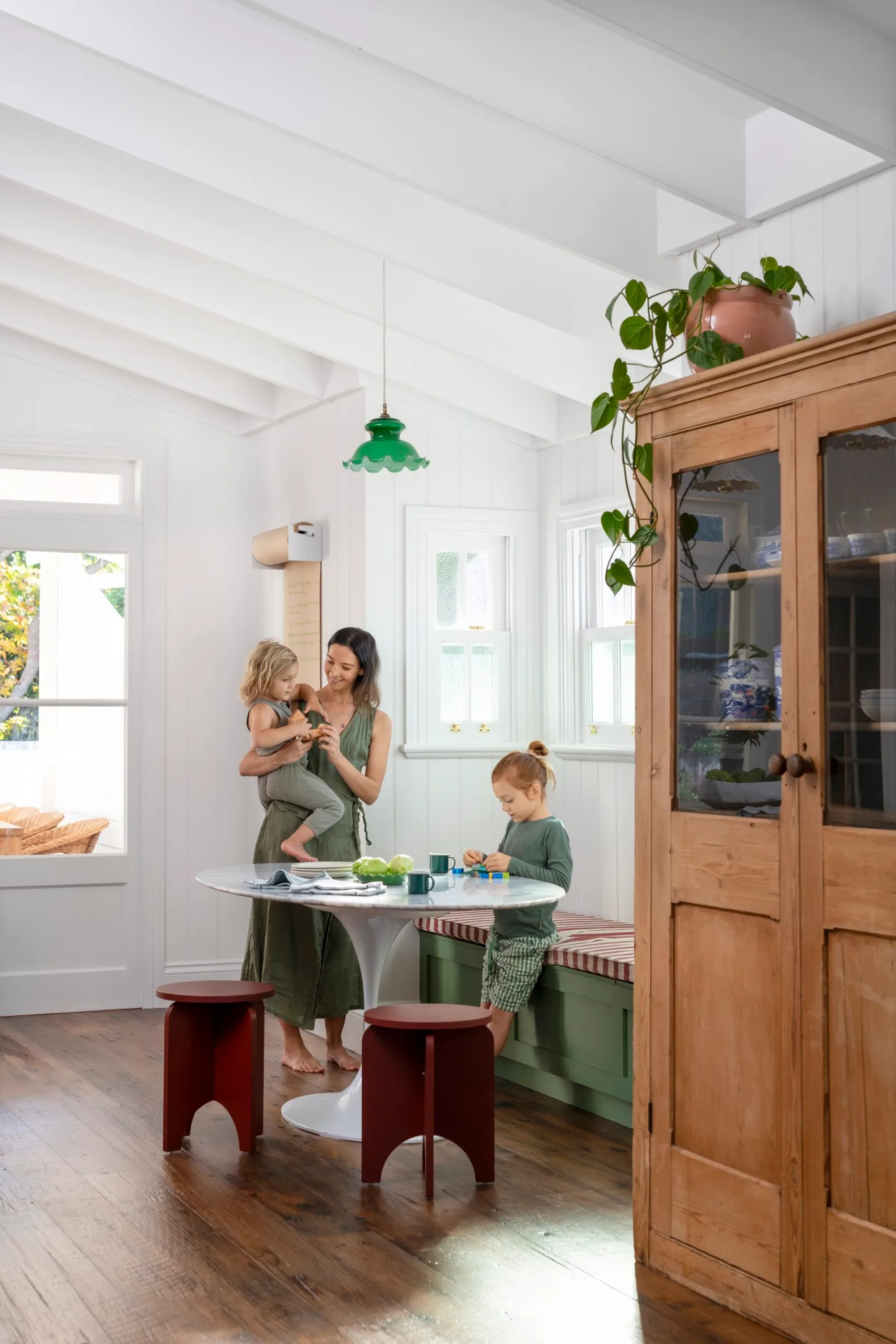 A mother with two children in a bright kitchen, with wooden floors, bench seating, and a table with green decor.