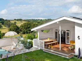 White house with wooden deck, outdoor dining table, small pool, umbrella, chairs, and scenic countryside view.