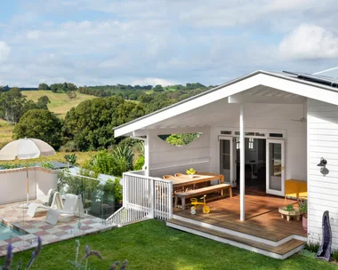 White house with wooden deck, outdoor dining table, small pool, umbrella, chairs, and scenic countryside view.
