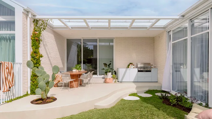 Outdoor kitchen with a cactus, round table, chairs, greenery, and a barbecue on a sunny day.