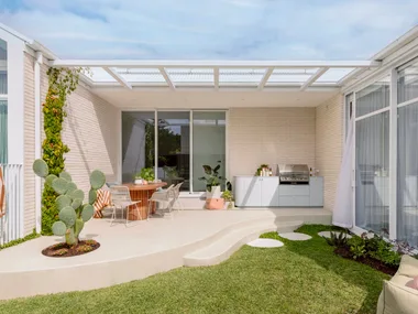 Outdoor kitchen with a cactus, round table, chairs, greenery, and a barbecue on a sunny day.