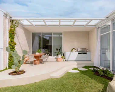 Outdoor kitchen with a cactus, round table, chairs, greenery, and a barbecue on a sunny day.