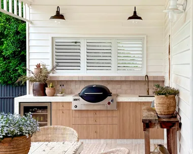 Outdoor kitchen with wooden cabinetry, a grill, white ceiling fan, and rustic dining table surrounded by wicker chairs.