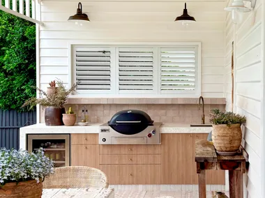 Outdoor kitchen with wooden cabinetry, a grill, white ceiling fan, and rustic dining table surrounded by wicker chairs.