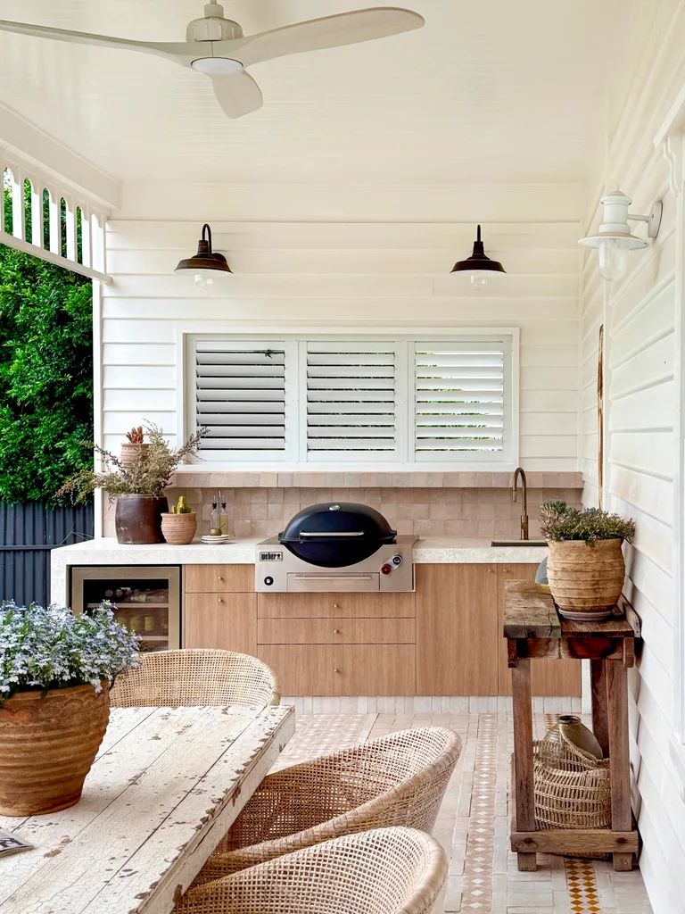 Outdoor kitchen with wooden cabinetry, a grill, white ceiling fan, and rustic dining table surrounded by wicker chairs.