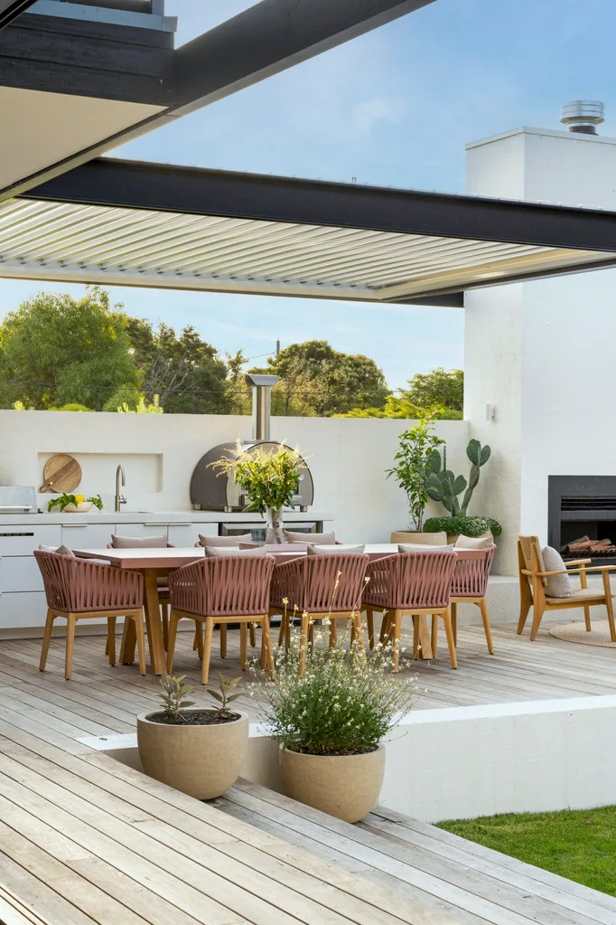 Outdoor kitchen with an alfresco dining area, pink chairs, plants, and a fireplace under a modern pergola.