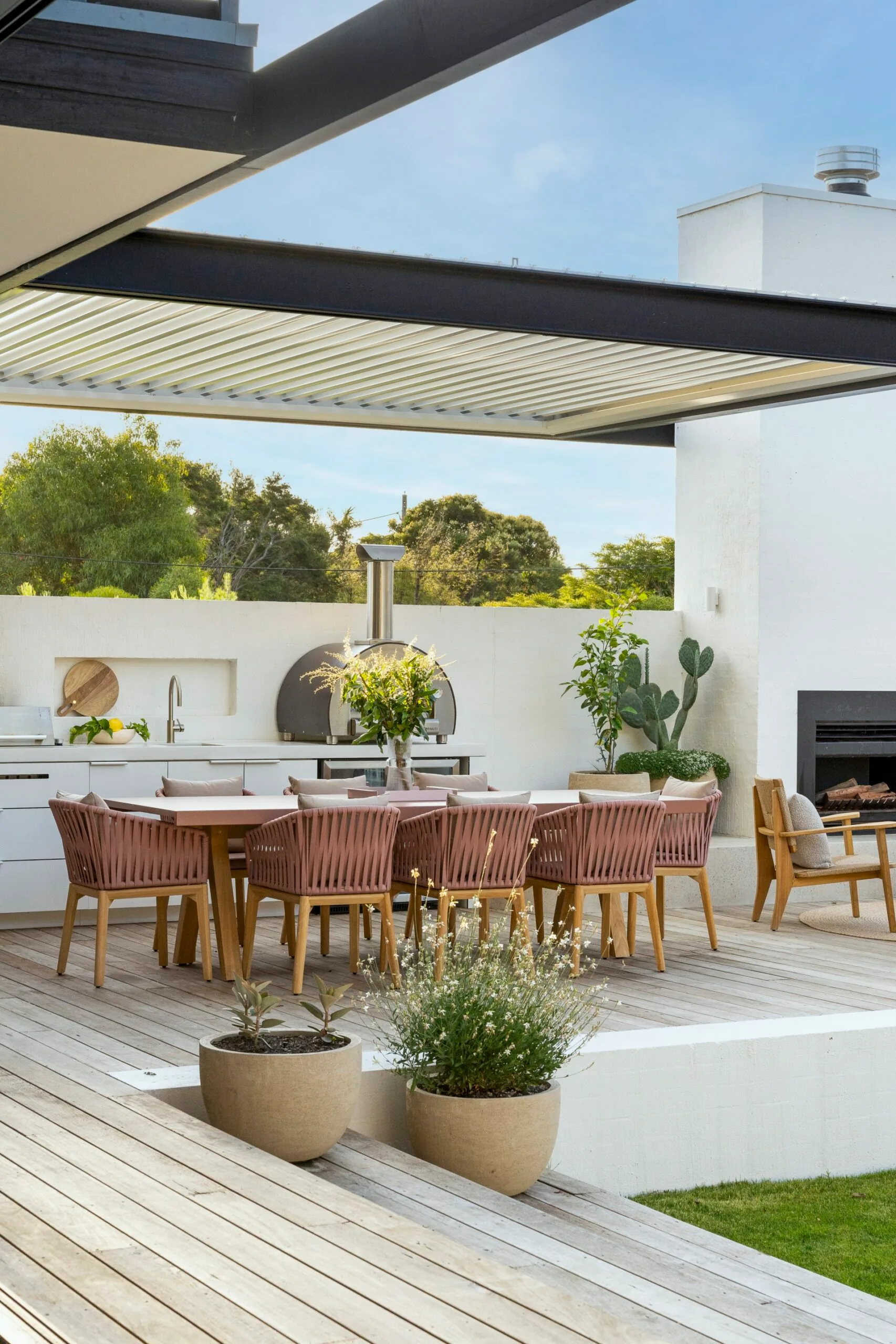 Outdoor kitchen with an alfresco dining area, pink chairs, plants, and a fireplace under a modern pergola.