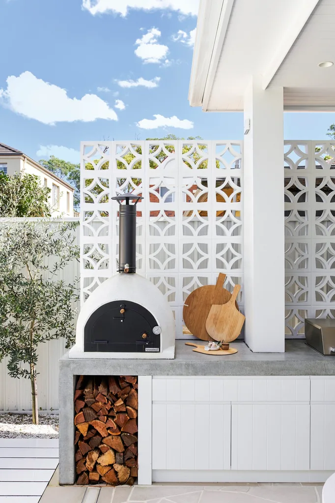 Outdoor kitchen with a pizza oven, stacked firewood, and wooden cutting boards, set against a decorative white panel.