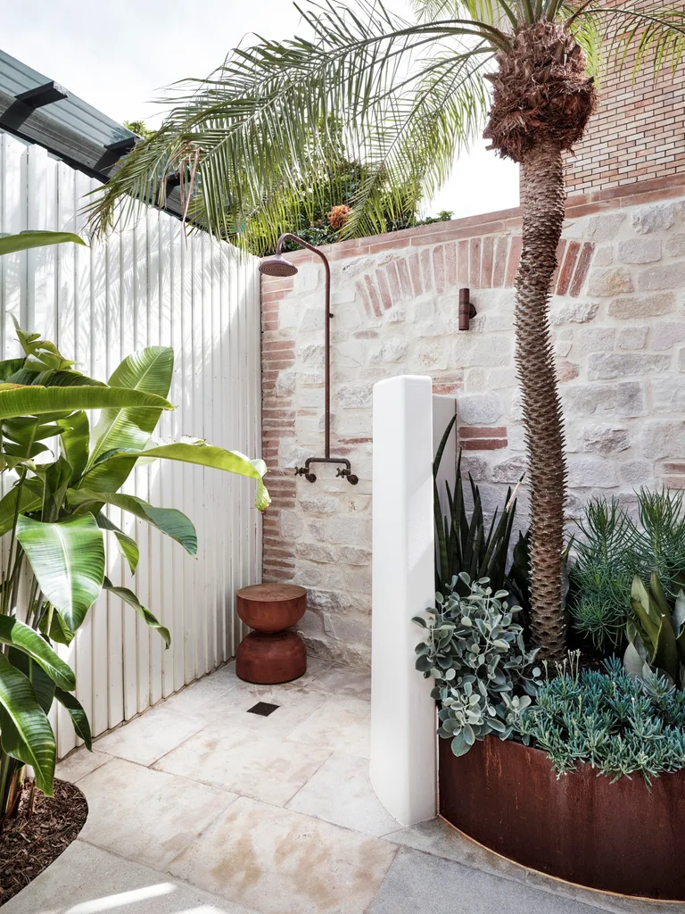 Outdoor shower with stone wall, palm tree, and greenery. Features a wooden stool and a rustic metal showerhead.