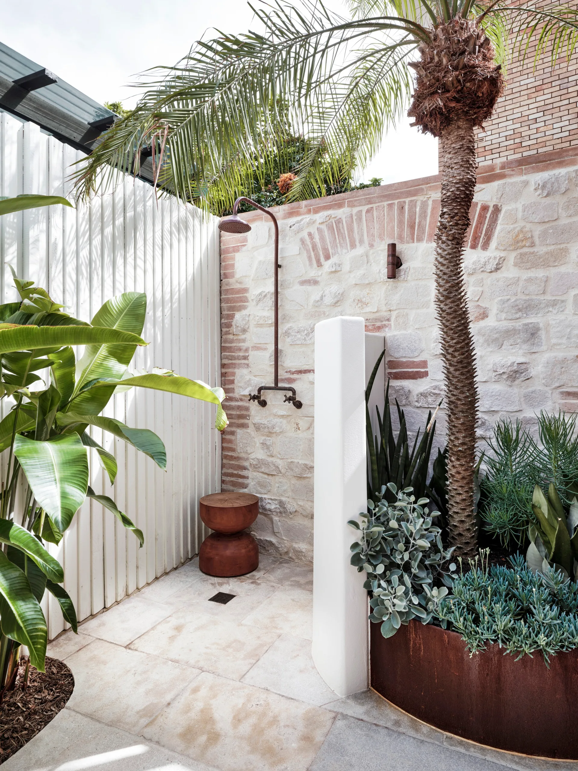 Outdoor shower with stone wall, palm tree, and greenery. Features a wooden stool and a rustic metal showerhead.