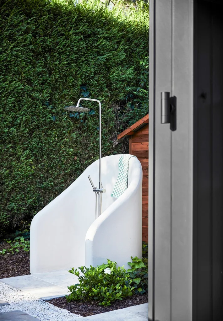 Outdoor shower area with a white chair-like enclosure, metal showerhead, green foliage background, and tiled flooring.