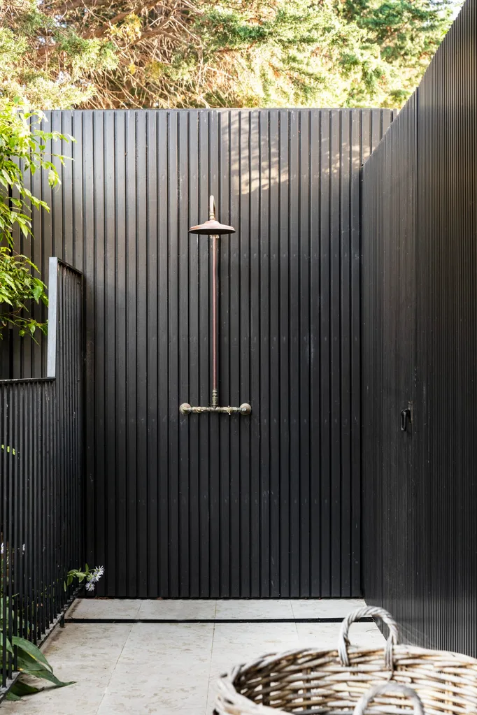 Outdoor shower with a wooden slat wall, surrounded by greenery and a wicker basket in the foreground.