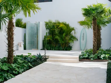 Outdoor shower area with tropical plants, surfboard, and palm trees in a bright courtyard setting.