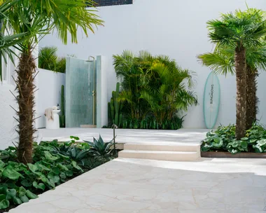 Outdoor shower area with tropical plants, surfboard, and palm trees in a bright courtyard setting.
