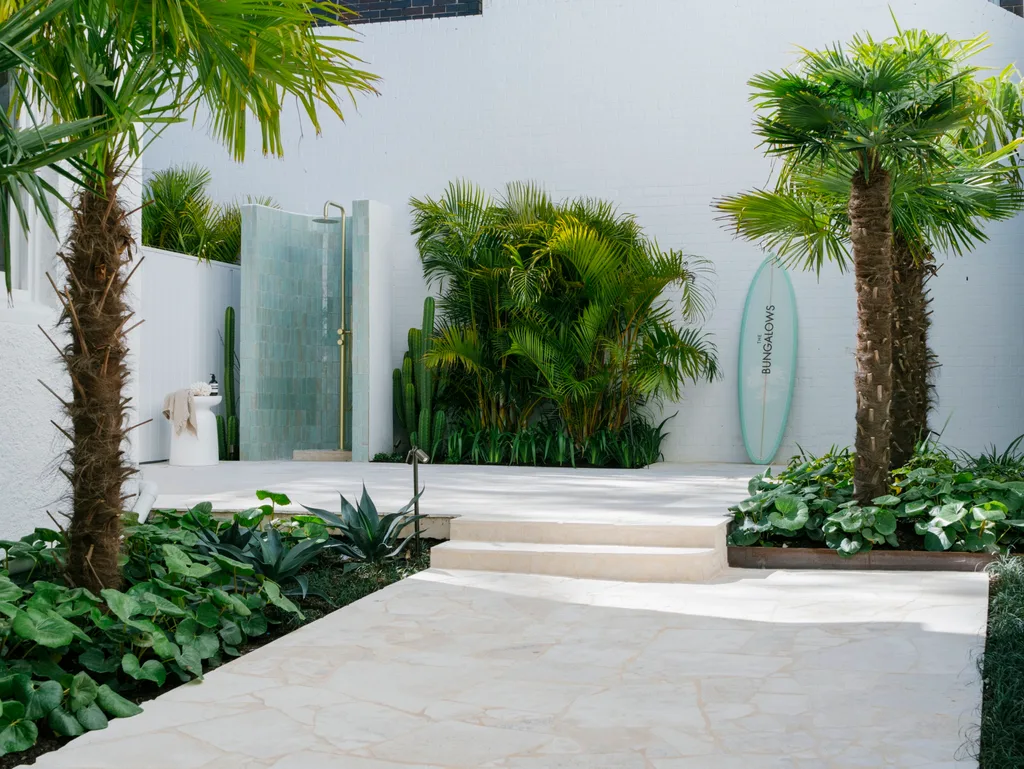 Outdoor shower area with tropical plants, surfboard, and palm trees in a bright courtyard setting.