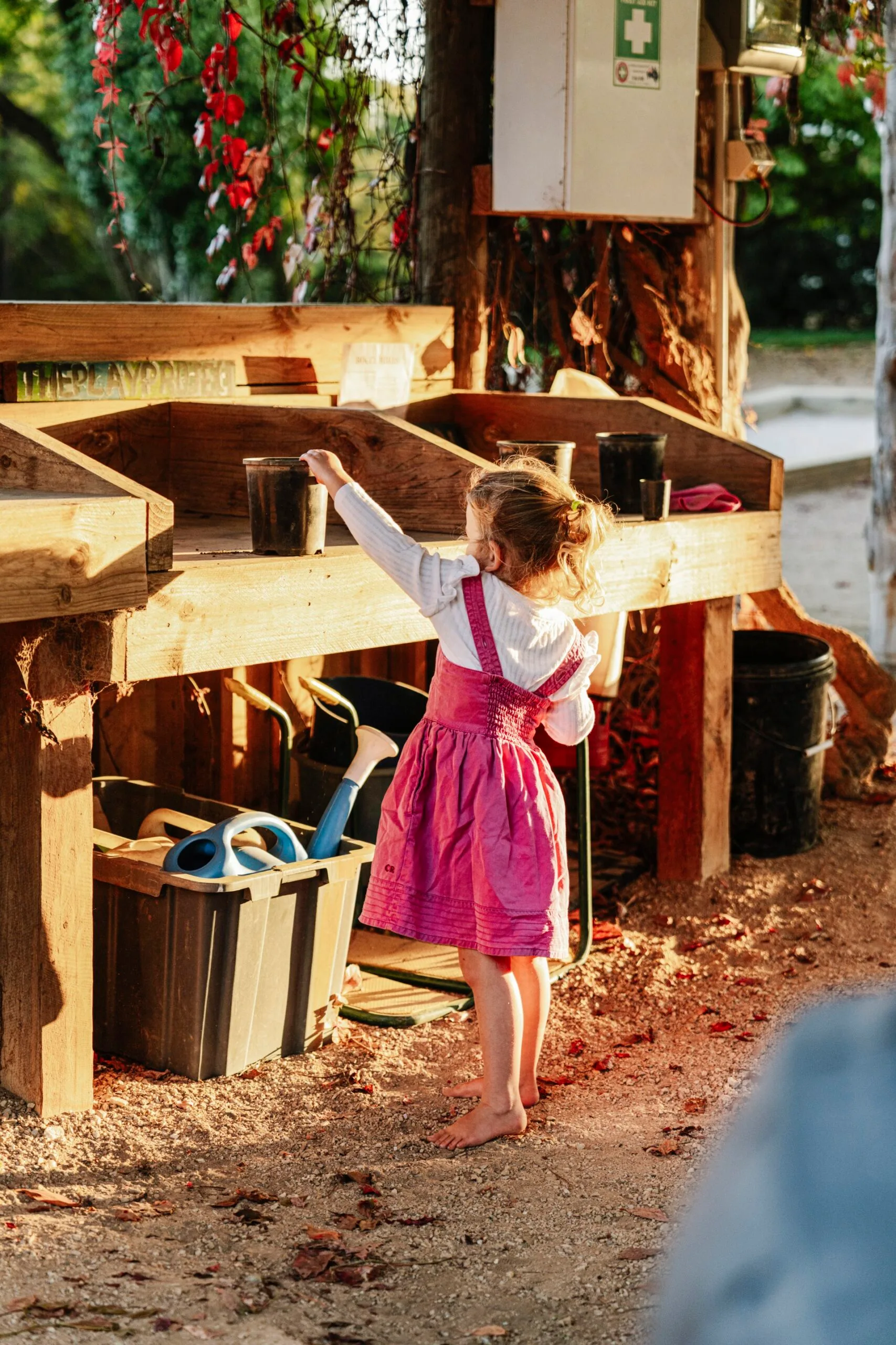 Young child in a pink dress plays at a wooden outdoor play station under the sun.