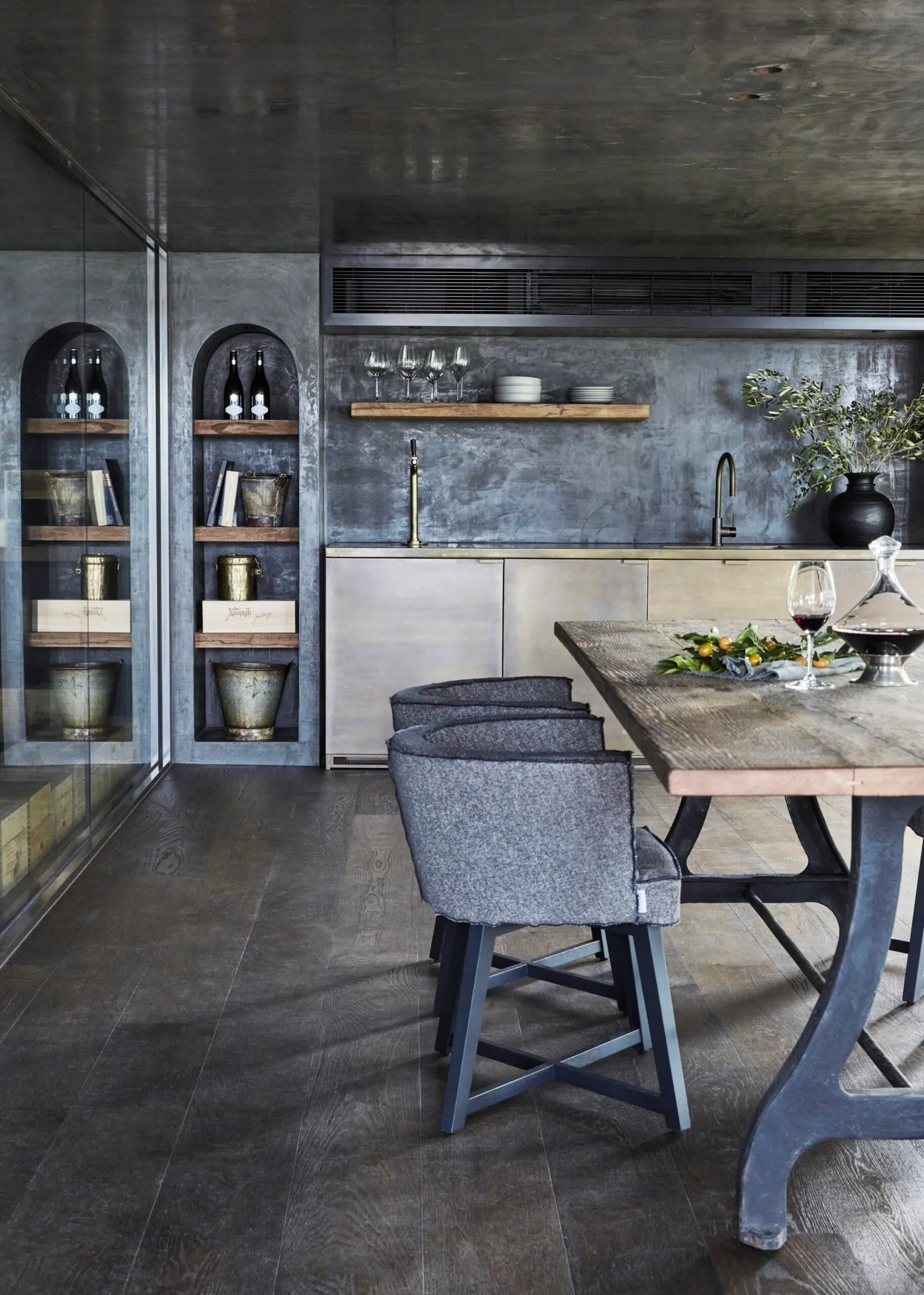 Modern kitchen or cocktail bar area with gray tones, wooden shelves, wine bottles, glasses, and a wooden table set for dining.