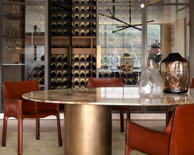 Dining room with a round marble table, leather chairs, modern chandelier, and a cellar in the background with glass doors.