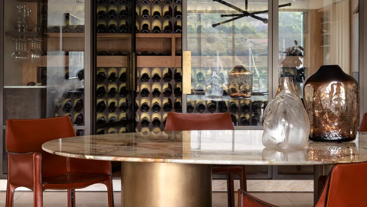 Dining room with a round marble table, leather chairs, modern chandelier, and a cellar in the background with glass doors.