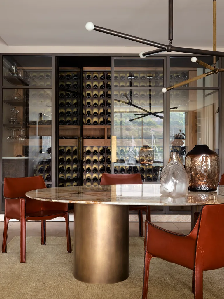 Dining room with a round marble table, leather chairs, modern chandelier, and a cellar in the background with glass doors.