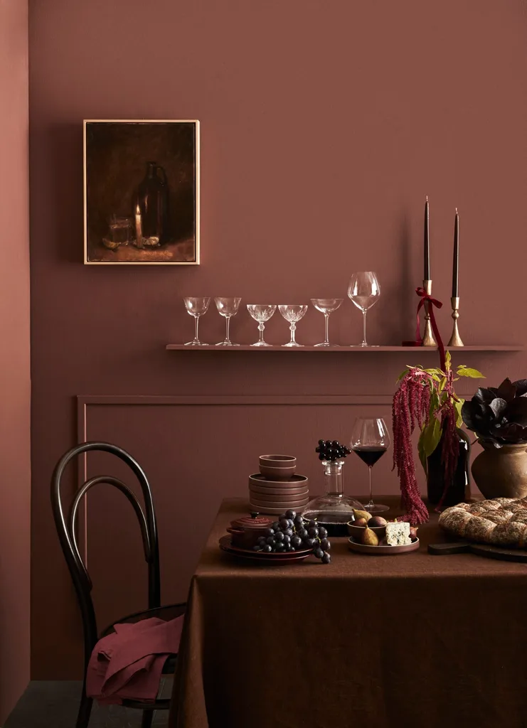 A moody dining setup with dark tableware, grapes, wine, and wall art, set against a deep maroon wall.