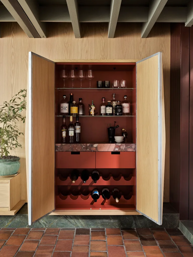 Open wooden cabinet with bottles, glasses, and wine rack, next to a potted plant, on brick tile floor.