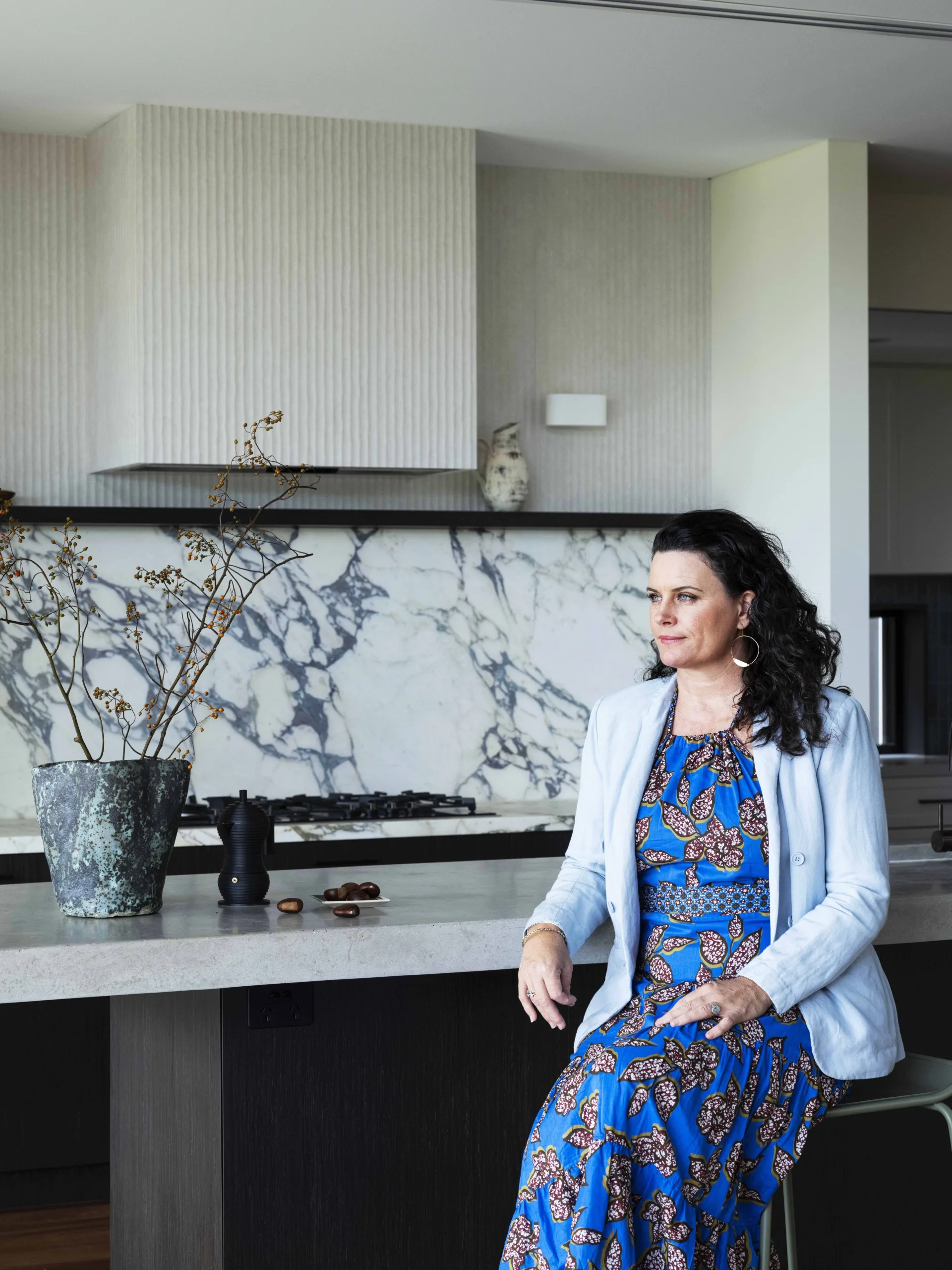 Woman in blue floral dress sitting on a kitchen stool, with a marble countertop and wall in the background.