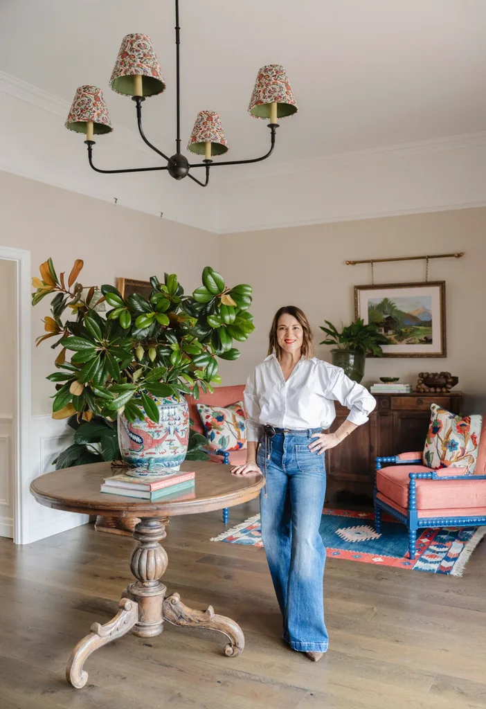 Woman in a white shirt and jeans stands by a round wooden table with a large potted plant in a stylish living room.