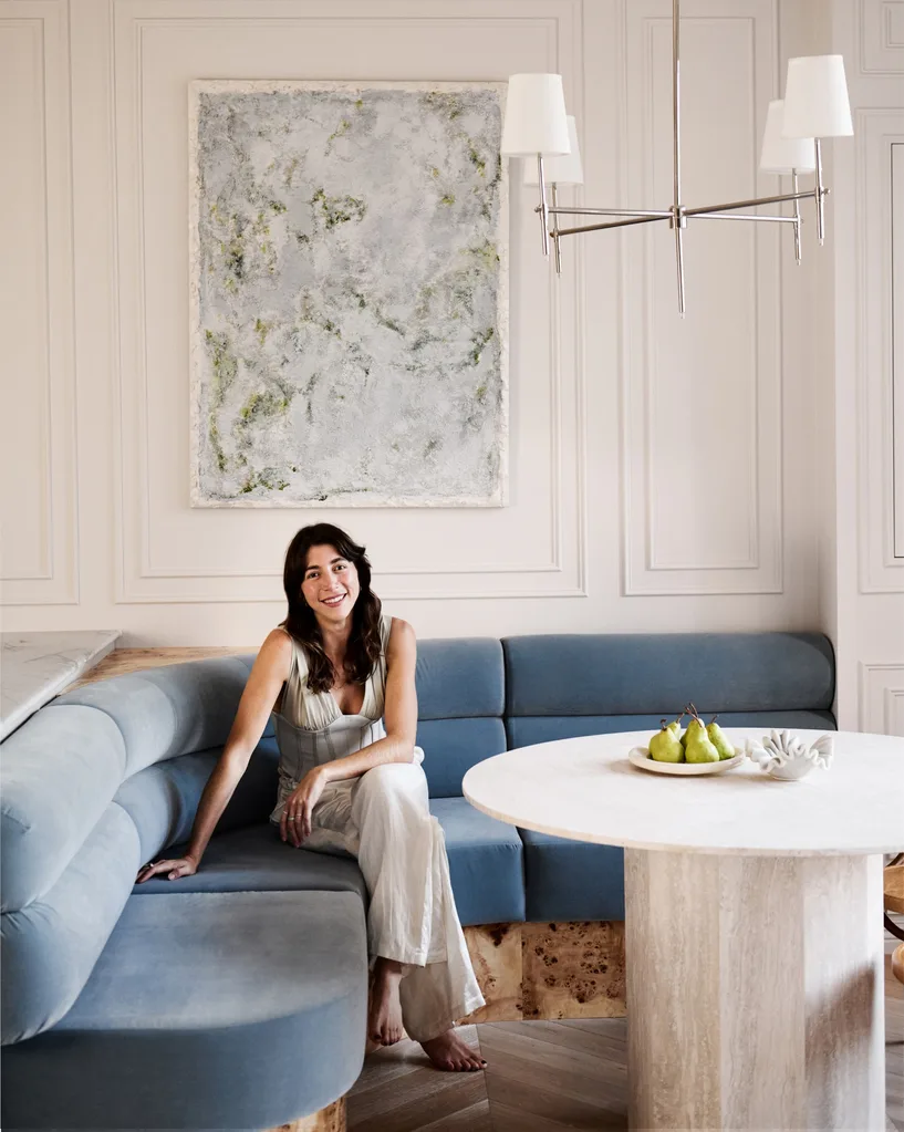 Australian designer seated on a blue couch in a stylish room with a marble table, artwork, and chandelier.