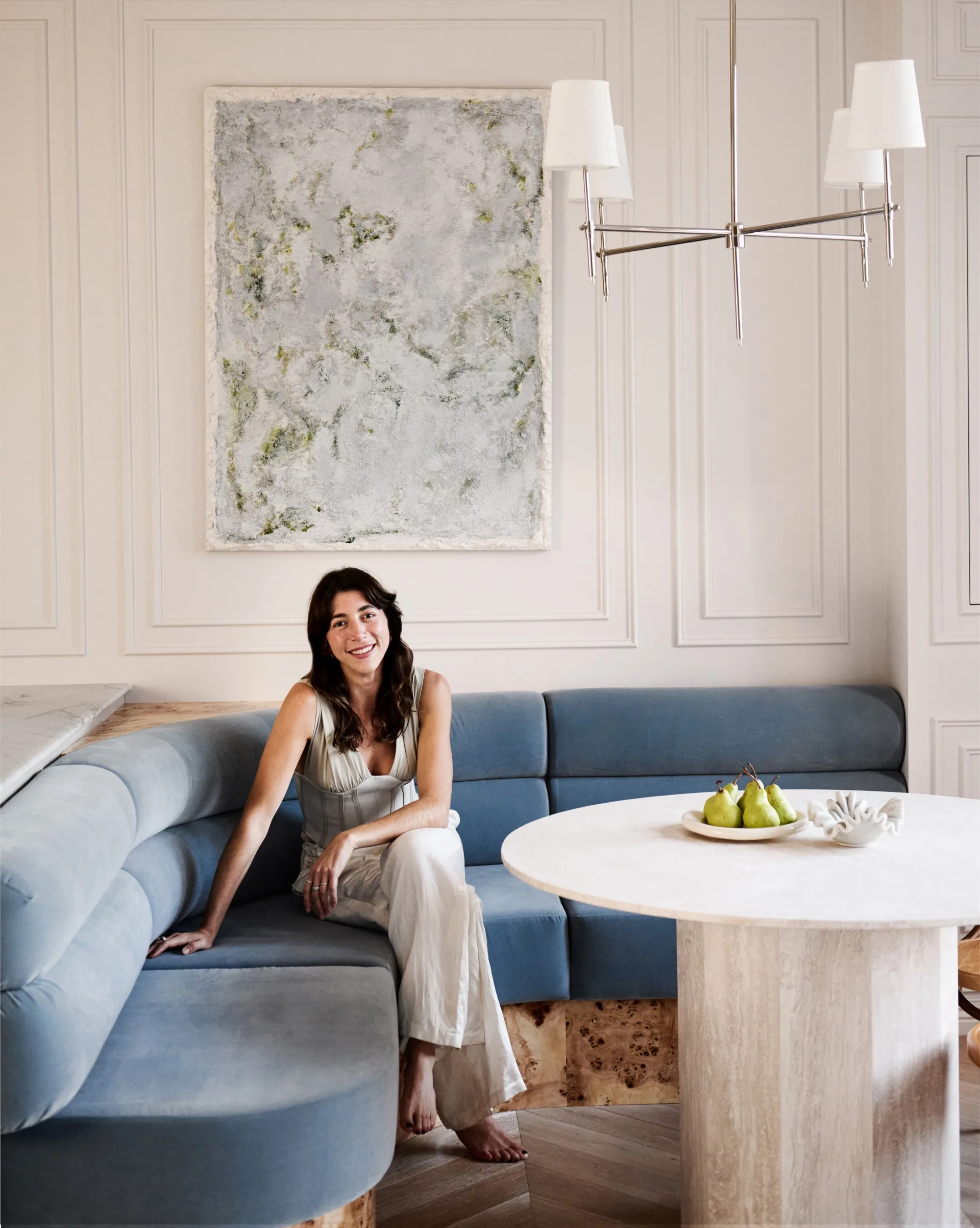 Australian designer seated on a blue couch in a stylish room with a marble table, artwork, and chandelier.