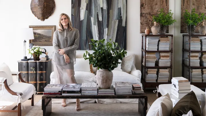 Woman standing in elegantly decorated living room with antique art, antique furniture, books, plants, and a turtle shell on the wall.