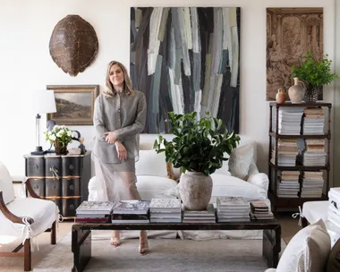 Woman standing in elegantly decorated living room with antique art, antique furniture, books, plants, and a turtle shell on the wall.