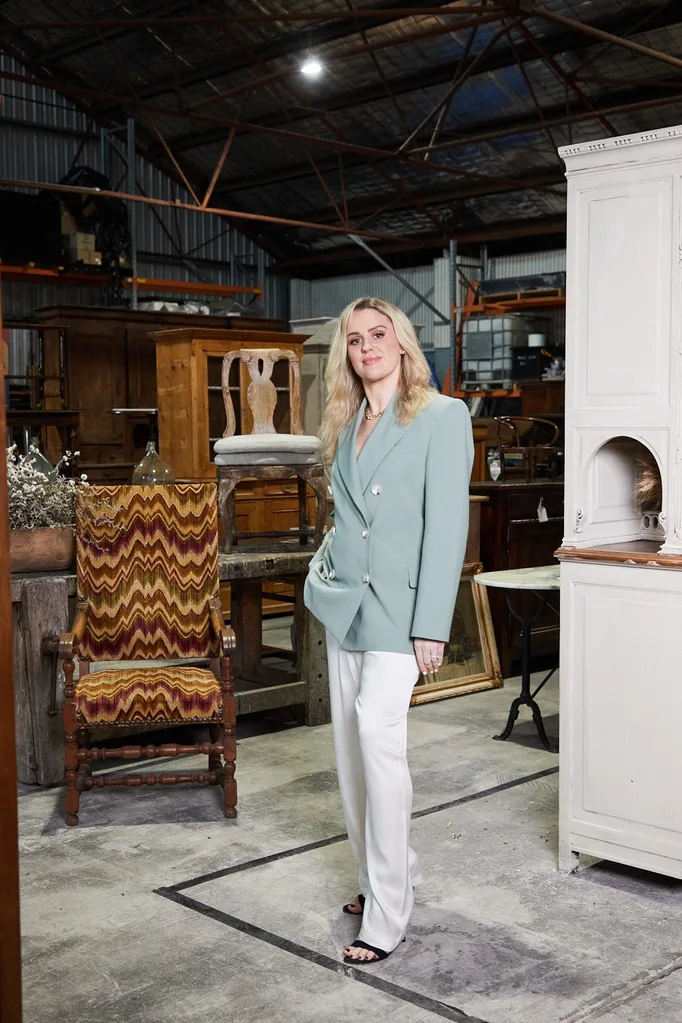 A woman in a light blue blazer stands in front of antique furniture, surrounded by vintage chairs and cabinets.