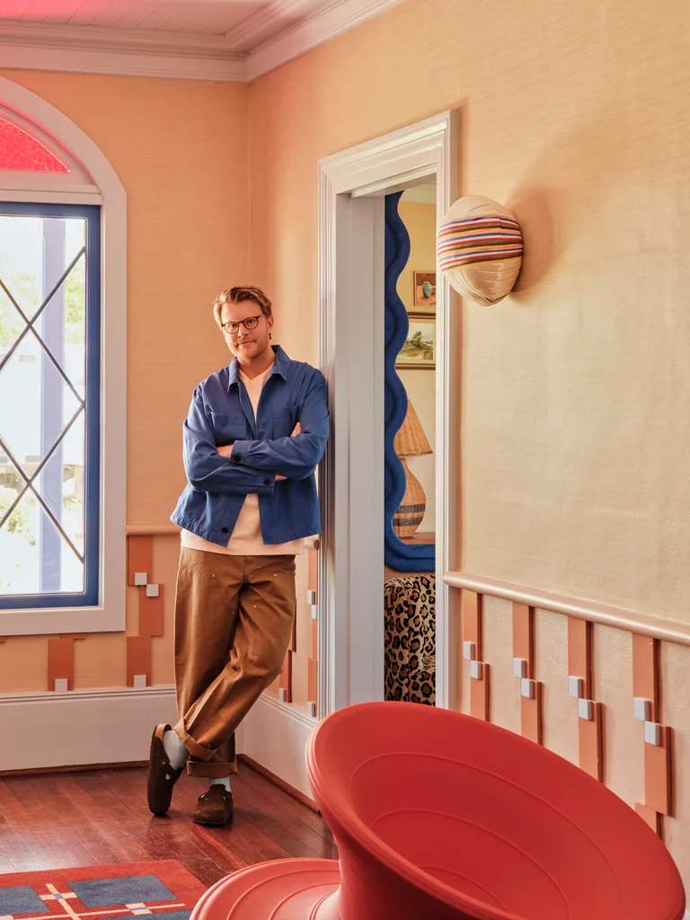 an Australian designer in a blue shirt and brown pants leans in a colorful, stylish room with modern decor.