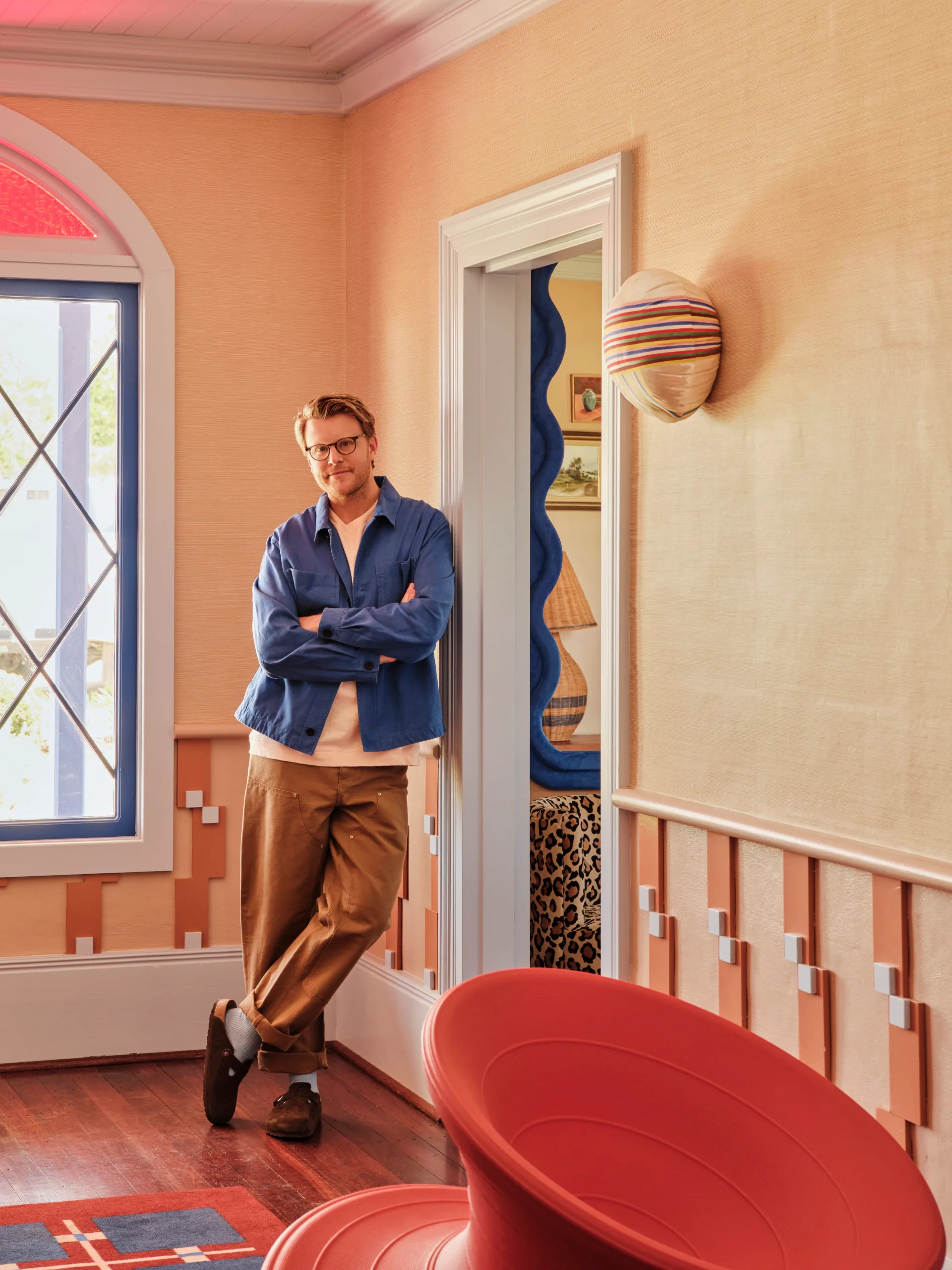 an Australian designer in a blue shirt and brown pants leans in a colorful, stylish room with modern decor.