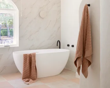 Modern bathroom with a freestanding white tub, beige towels, and marble tiles. Natural light from a nearby window.