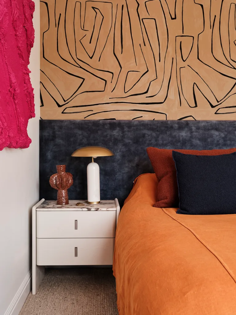 Bedroom with orange bedding, abstract wall art, white nightstand, decorative lamp, and textured pink wall piece. It's the main bedroom in a family home.