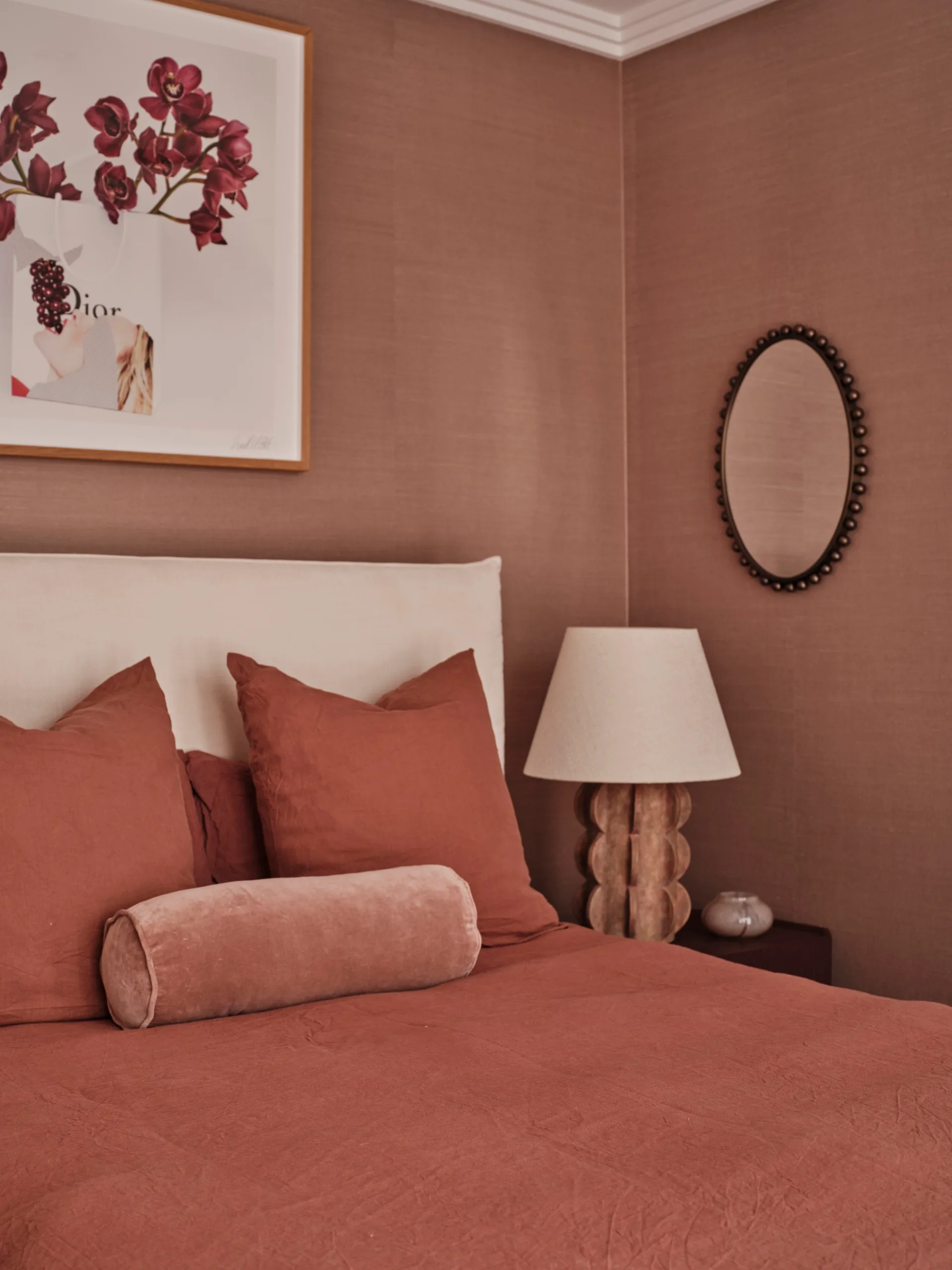 Bedroom corner with terracotta bedding which matches the terracotta coloured wallpaper, round pillow, modern lamp, oval mirror, and floral artwork on the wall.